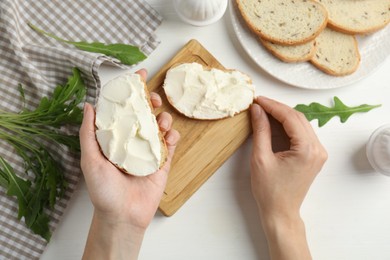Woman holding bread with cream cheese at white wooden table, closeup