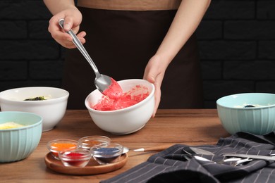 Photo of Woman mixing cream with red food coloring at wooden table, closeup