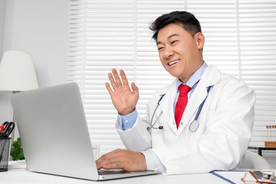 Happy doctor with laptop consulting patient at white desk in clinic. Online medicine