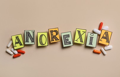 Word Anorexia made of papers with letters near pills on beige background, flat lay