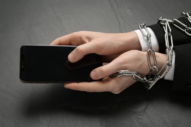 Photo of Man holding smartphone in chained hands at black table, closeup. Internet addiction