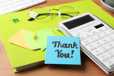 Light blue paper note with phrase Thank You, calculator and notebook on wooden table