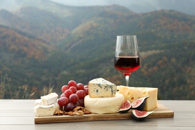 Different types of delicious cheeses, fruits and wine on wooden table against mountain landscape
