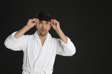 Surprised young man in bathrobe and eye sleeping mask on black background. Space for text