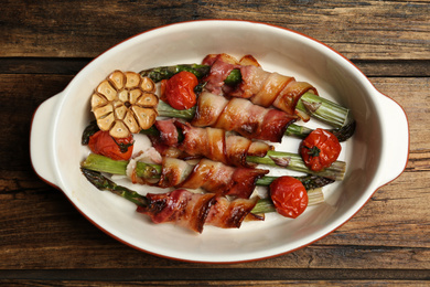 Oven baked asparagus wrapped with bacon in ceramic dish on wooden table, top view