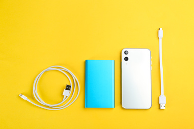 Mobile phone, portable charger and cables on yellow background, flat lay
