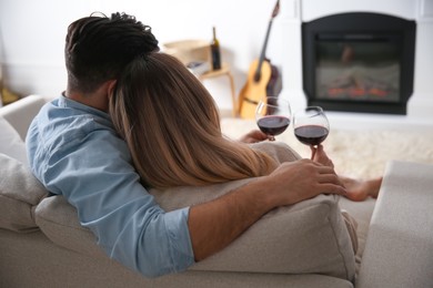 Couple with glasses of wine resting on sofa near fireplace at home, back view