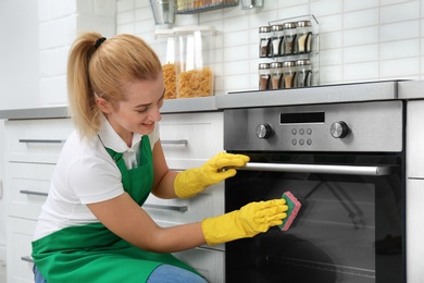 Female janitor cleaning oven with sponge in kitchen