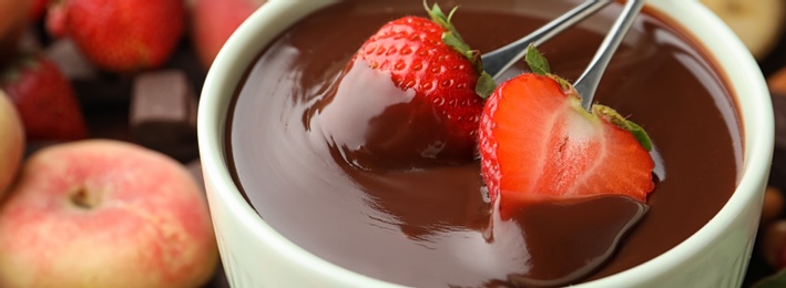 Photo of Fondue forks with strawberries in bowl of melted chocolate on table, closeup