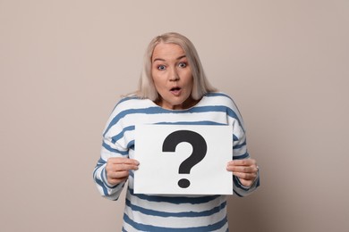 Emotional mature woman holding paper with question mark on beige background