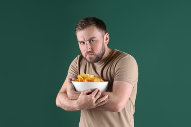 Greedy young man hiding bowl with chips on green background