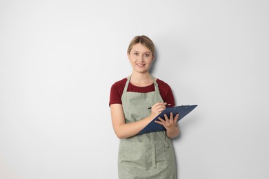 Beautiful young woman in clean apron with clipboard on light grey background