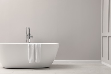 Modern ceramic bathtub with towel near light wall indoors, space for text