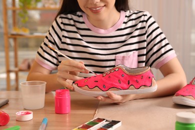 Woman painting on sneaker at wooden table indoors, closeup. Customized shoes