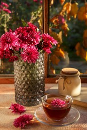 Photo of Beautiful chrysanthemum flowers in vase and cup of tea on beige textured table near window. Autumn still life