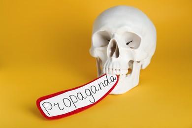 Photo of Information warfare concept. Human skull and paper card with word Propaganda in teeth on yellow background