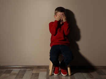 Photo of Scared little boy on wooden chair near beige wall, space for text. Domestic violence concept