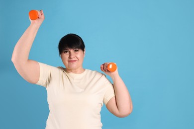 Happy overweight mature woman doing exercise with dumbbells on light blue background, space for text