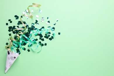 Shiny confetti and streamers with party cracker on light green background, top view. Space for text