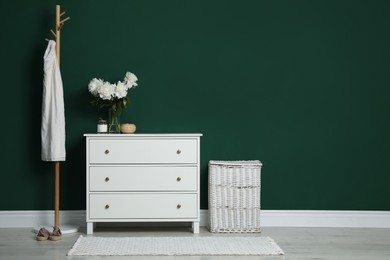 Modern white chest of drawers, coat stand and wicker basket near green wall indoors. Space for text