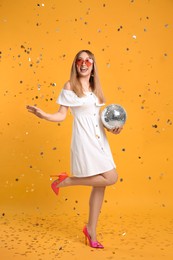 Happy young woman with disco ball and confetti on yellow background