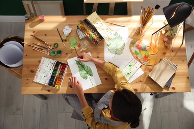 Young woman drawing leaf with watercolors at table indoors, top view