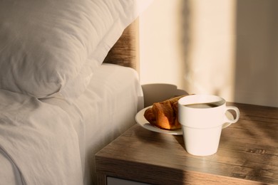 Cup of coffee and croissant on night stand near bed in morning