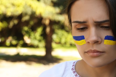 Photo of Sad young woman with drawings of Ukrainian flag on face outdoors, closeup. Space for text