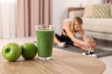 Photo of Young woman in fitness clothes doing exercise at home, focus on glass of smoothie and apples