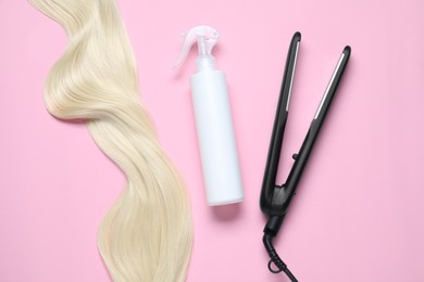 Photo of Spray bottle with thermal protection, iron and lock of blonde hair on pink background, flat lay