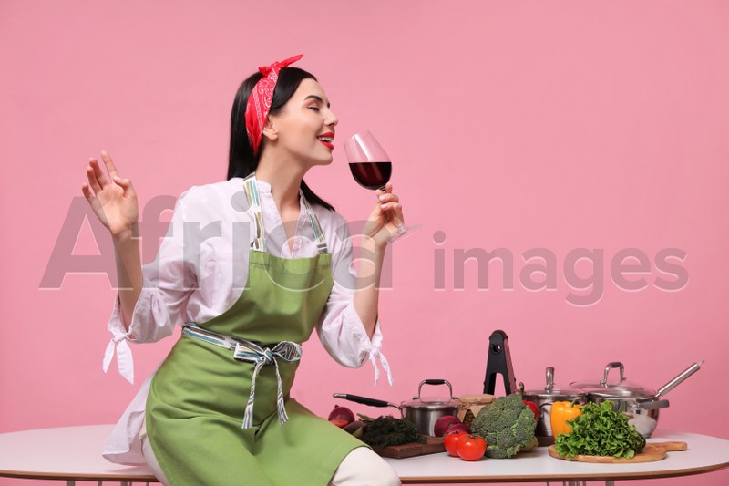 Young housewife with glass of wine, vegetables and different utensils on pink background. Space for text