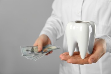 Dentist holding ceramic model of tooth and dollar banknotes on grey background, closeup. Expensive treatment