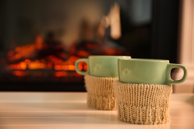 Cups with hot drink on light table against fireplace, space for text
