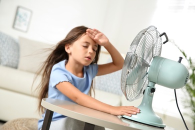 Little girl suffering from heat in front of fan at home. Summer season