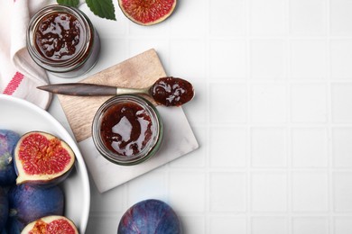 Photo of Glass jars of tasty sweet fig jam and fruits on white tiled table, flat lay. Space for text