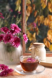 Beautiful chrysanthemum flowers in vase and cup of tea on beige textured table near window. Autumn still life