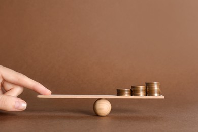Woman balancing miniature seesaw with stacks of coins on brown background, closeup. Harmony concept