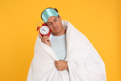 Man in sleeping mask wrapped with blanket holding alarm clock on yellow background