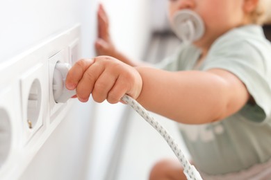 Cute baby playing with plug and electrical socket at home, closeup. Dangerous situation