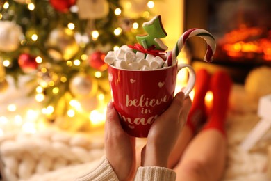Photo of Woman holding cup of sweet drink near fireplace at home, closeup. Cozy winter holidays atmosphere