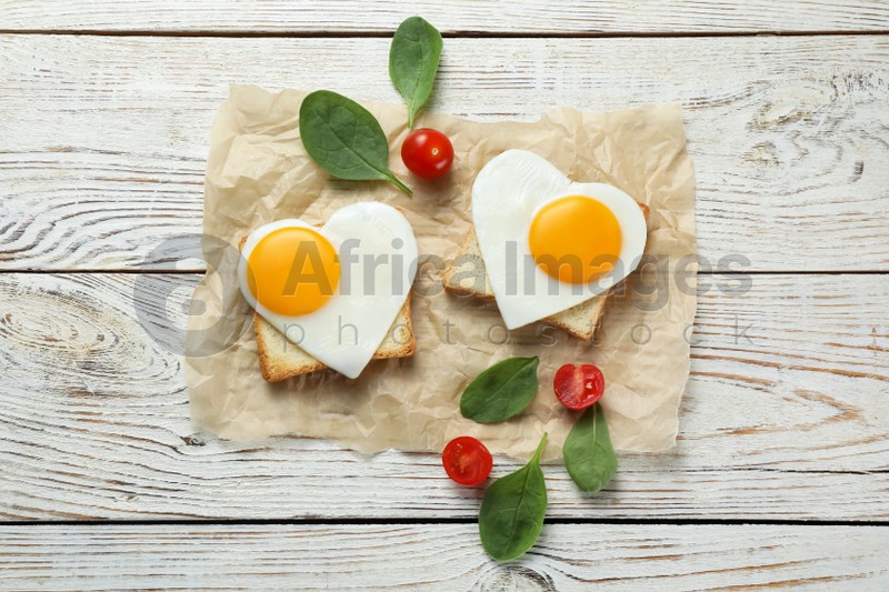 Heart shaped fried eggs with toasts, spinach and tomatoes on white wooden table, flat lay