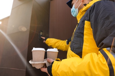 Courier in medical mask holding takeaway drinks and ringing doorbell outdoors, focus on paper cups. Delivery service during quarantine due to Covid-19 outbreak