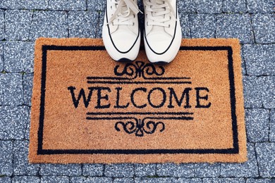 Photo of Doormat with word Welcome and stylish sneakers on pavement outdoors, flat lay