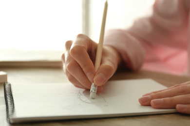 Woman correcting picture in notepad with pencil eraser at wooden table, closeup