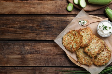 Delicious zucchini fritters with sour cream served on wooden table, flat lay. Space for text