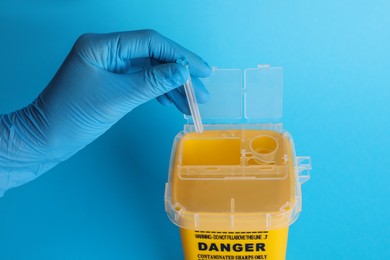 Doctor throwing used syringe needle into sharps container on light blue background, closeup