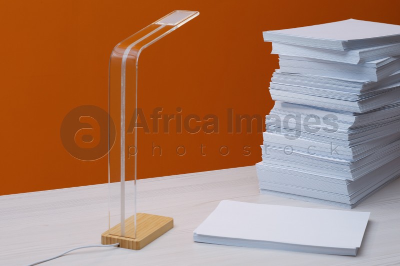 Stack of paper sheets and lamp on white wooden table near orange wall