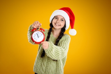 Girl in Santa hat with alarm clock on yellow background. New Year countdown