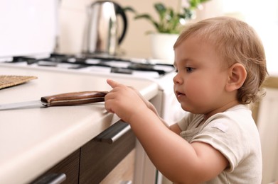 Photo of Little child touching sharp knife indoors. Dangers in kitchen