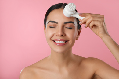 Young woman using facial cleansing brush on pink background. Washing accessory